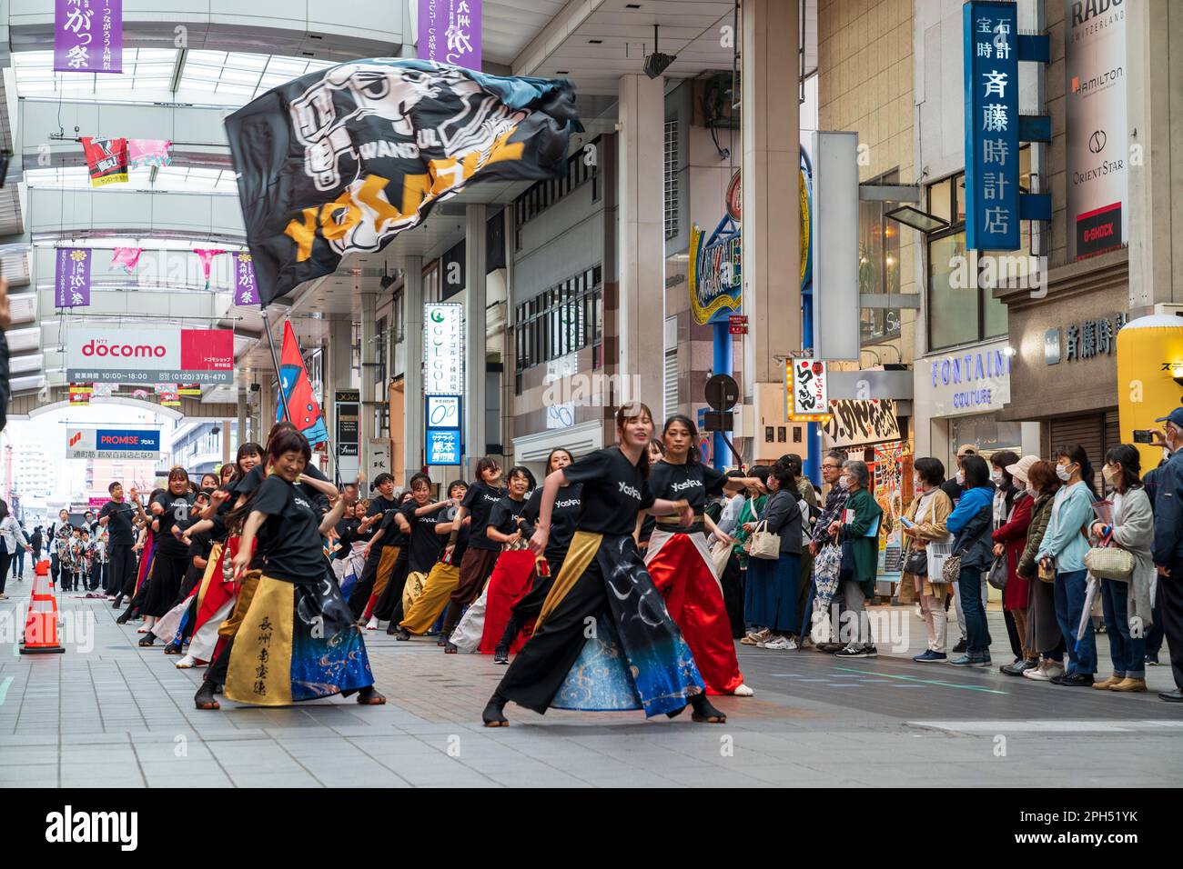 Team of young adult Japanese Yosakoi dancers wearing colourful yukata tunics while dancing in the middle of a covered shopping street, Sunroad Shinshigai, in Kumamoto during the annual springtime Kyusyu gassai dance festival. Some spectators watching. Stock Photo