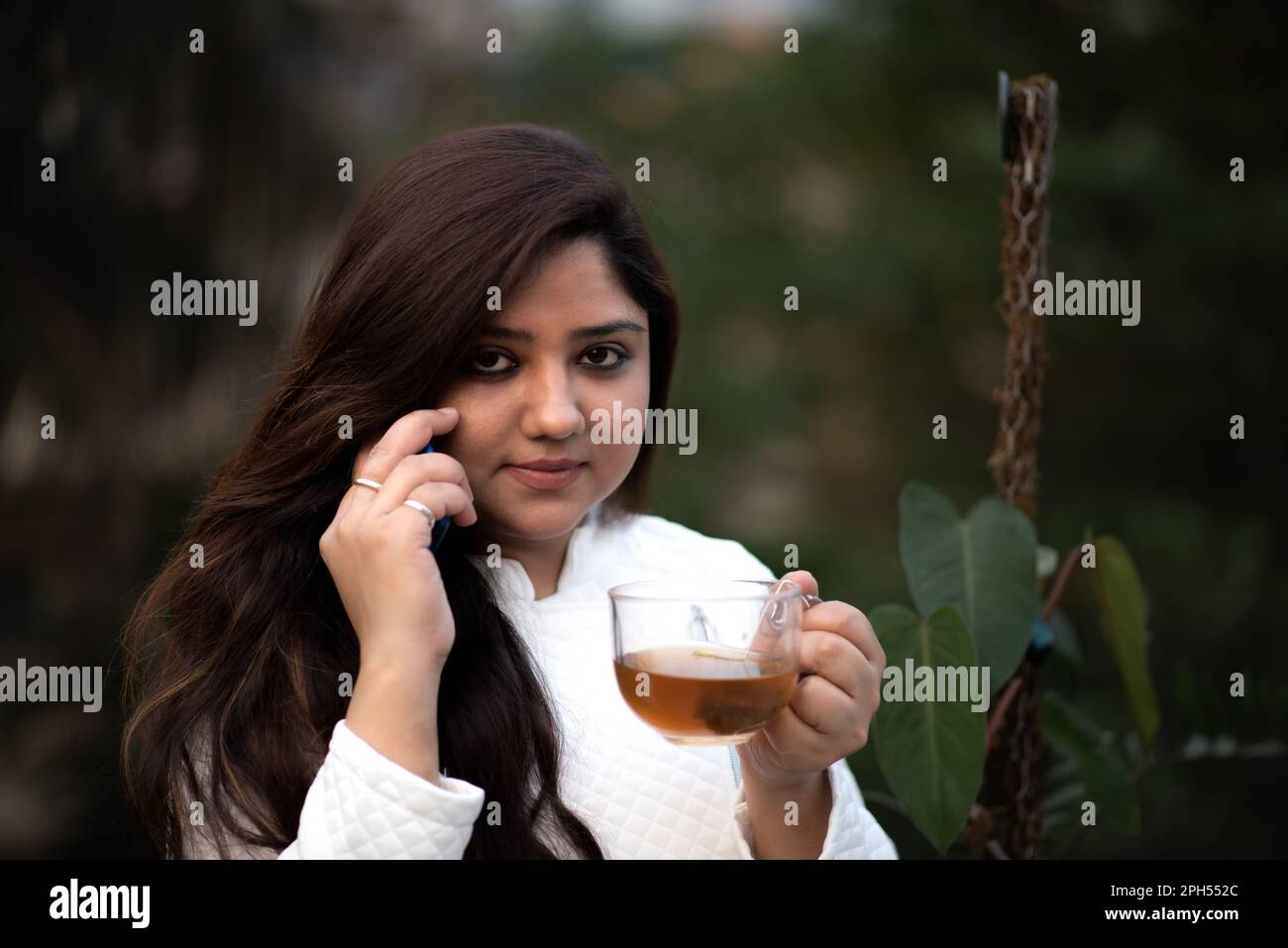 Women is on call on mobile phone holding black tea in a transparent tea cup wearing white woollen jacket in garden Stock Photo
