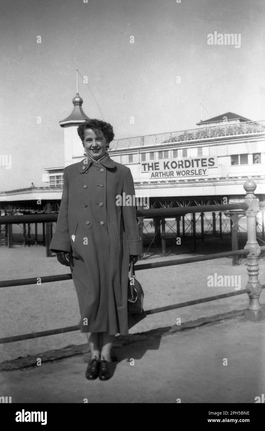 1954, historical, a lady holidaymaker in a long coat standing for a photo infront of the Central pier at the coastal town of Blackpool, Lancashire, England, UK. Two acts appearing there that year were the The Kordites and Arthur Worsley, whose names are written on the side of the pier building.  Arthur Worsley was a popular British ventriloquist of the era with regular TV appearances. The Kordites were a male and female vocal group, who also performed sketches with songs and were a regular act on the BBC light programme, 'Variety Fanfare'. Stock Photo
