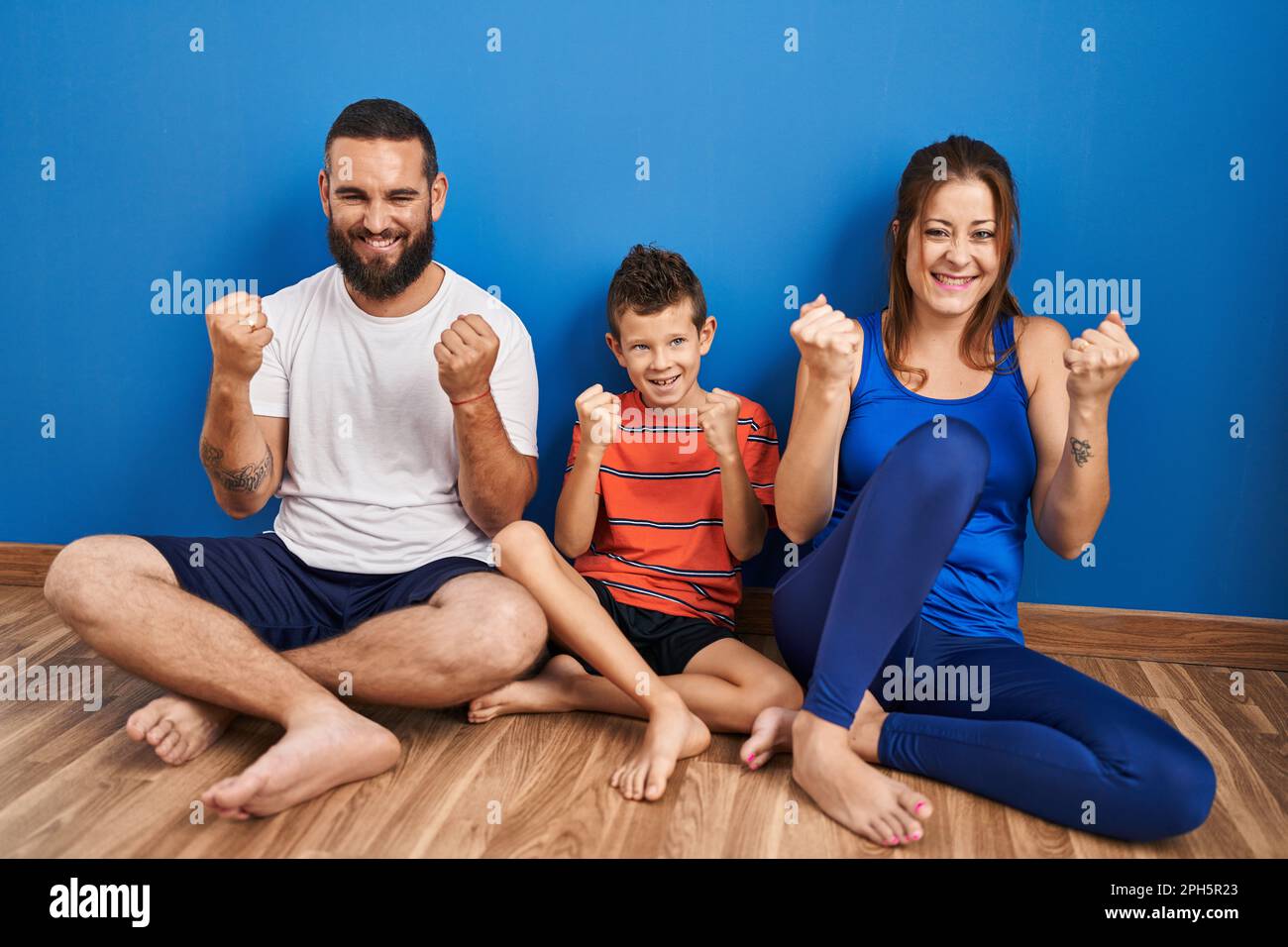 Family of three sitting on the floor at home very happy and excited doing winner gesture with arms raised, smiling and screaming for success. celebrat Stock Photo