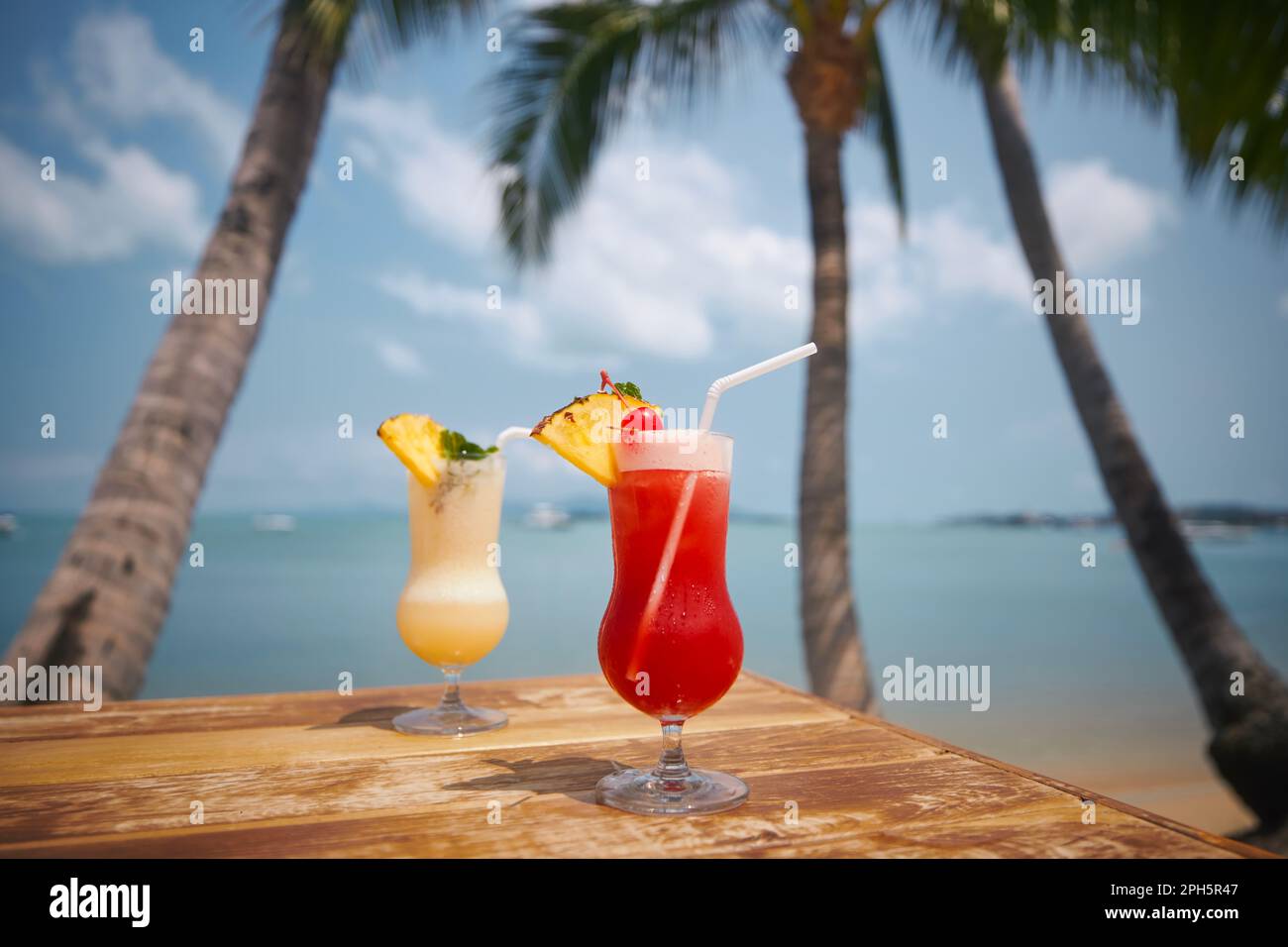Two cocktails under palm trees on beautiful beach. Singapore Sling and Pina Colada drinks on wooden table. Stock Photo