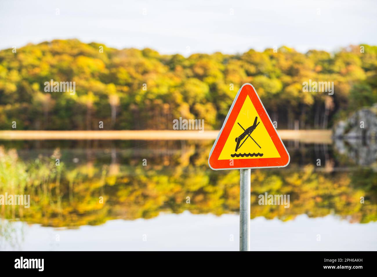 A bright yellow triangle-shaped warning sign prohibiting diving in a peaceful Swedish lake, emphasizing the importance of communication. Stock Photo