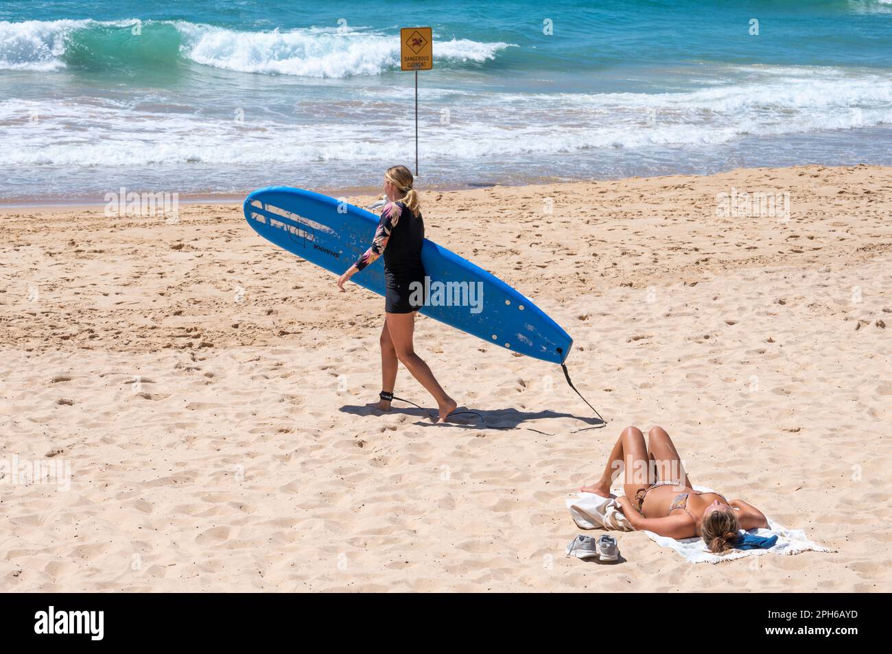As a sun bather soaks up the rays, a surfer carries her board along the beach at Manly, Sydney, New South Wales, Australia against a backdrop of the b Stock Photo