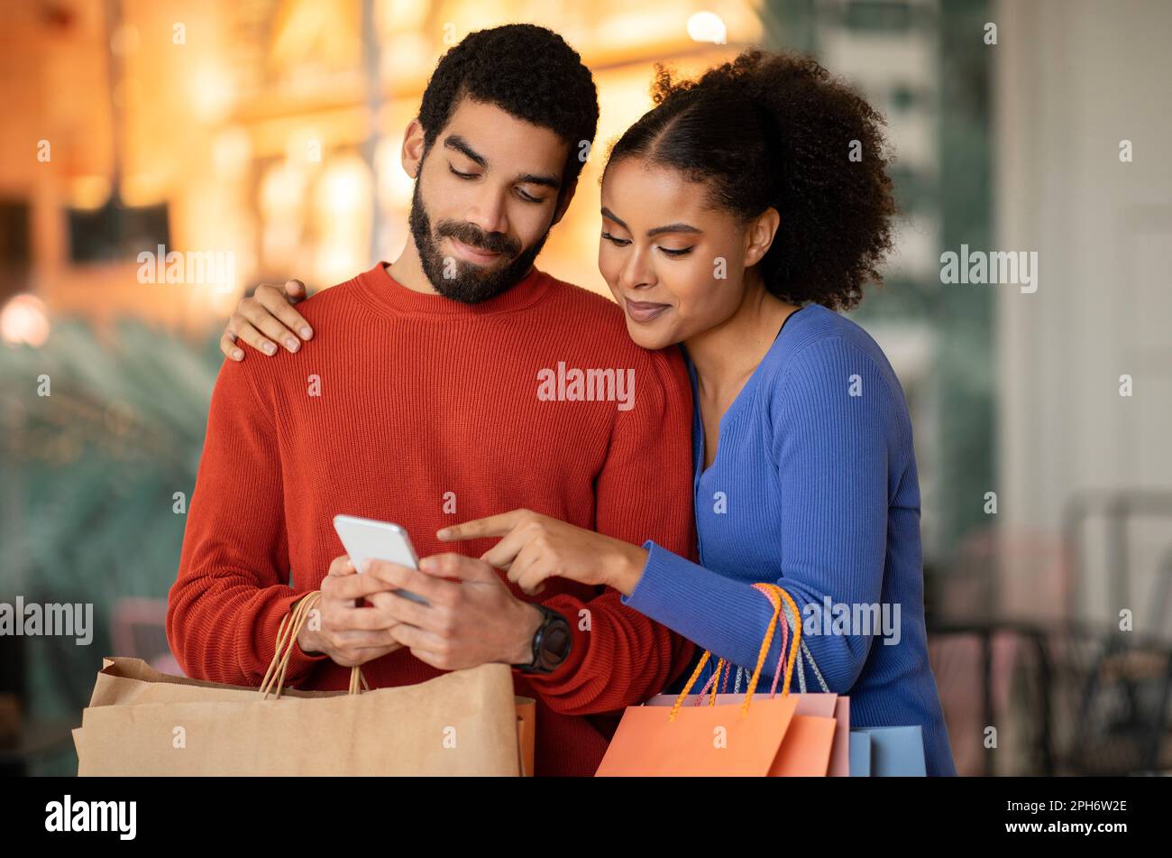 Multiethnic Spouses Shopping Using Cellphone Pointing Finger Standing At Mall Stock Photo