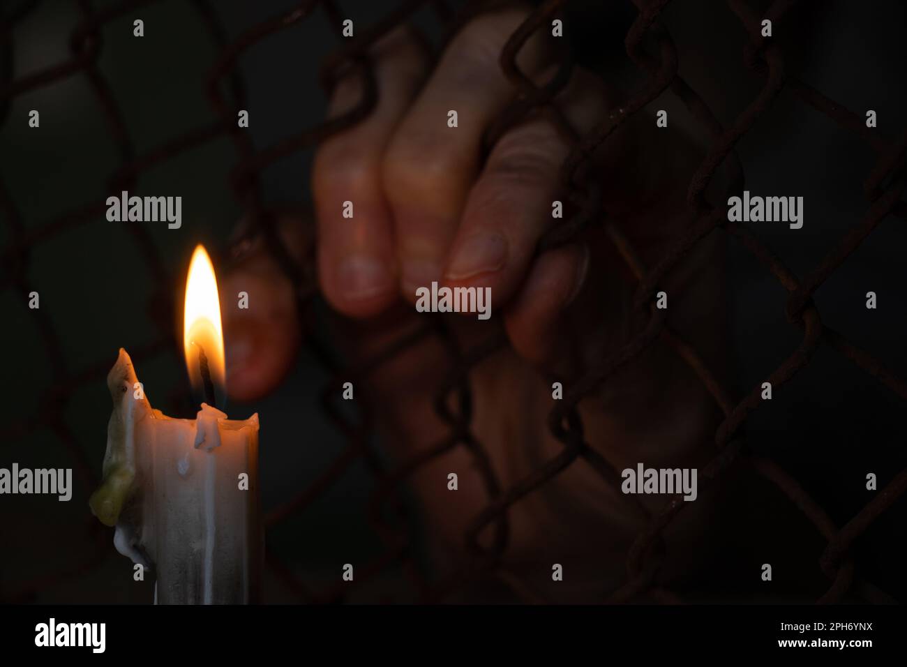 old female hands hang on a fence made of iron mesh in the lock on the background of a burning candle Stock Photo