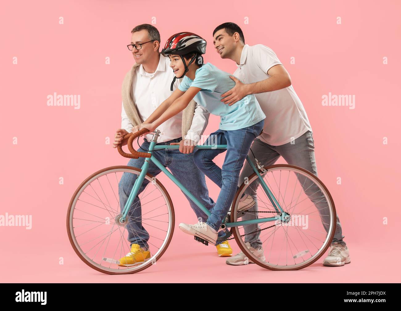 Young man with his father teaching little boy to ride bike on pink