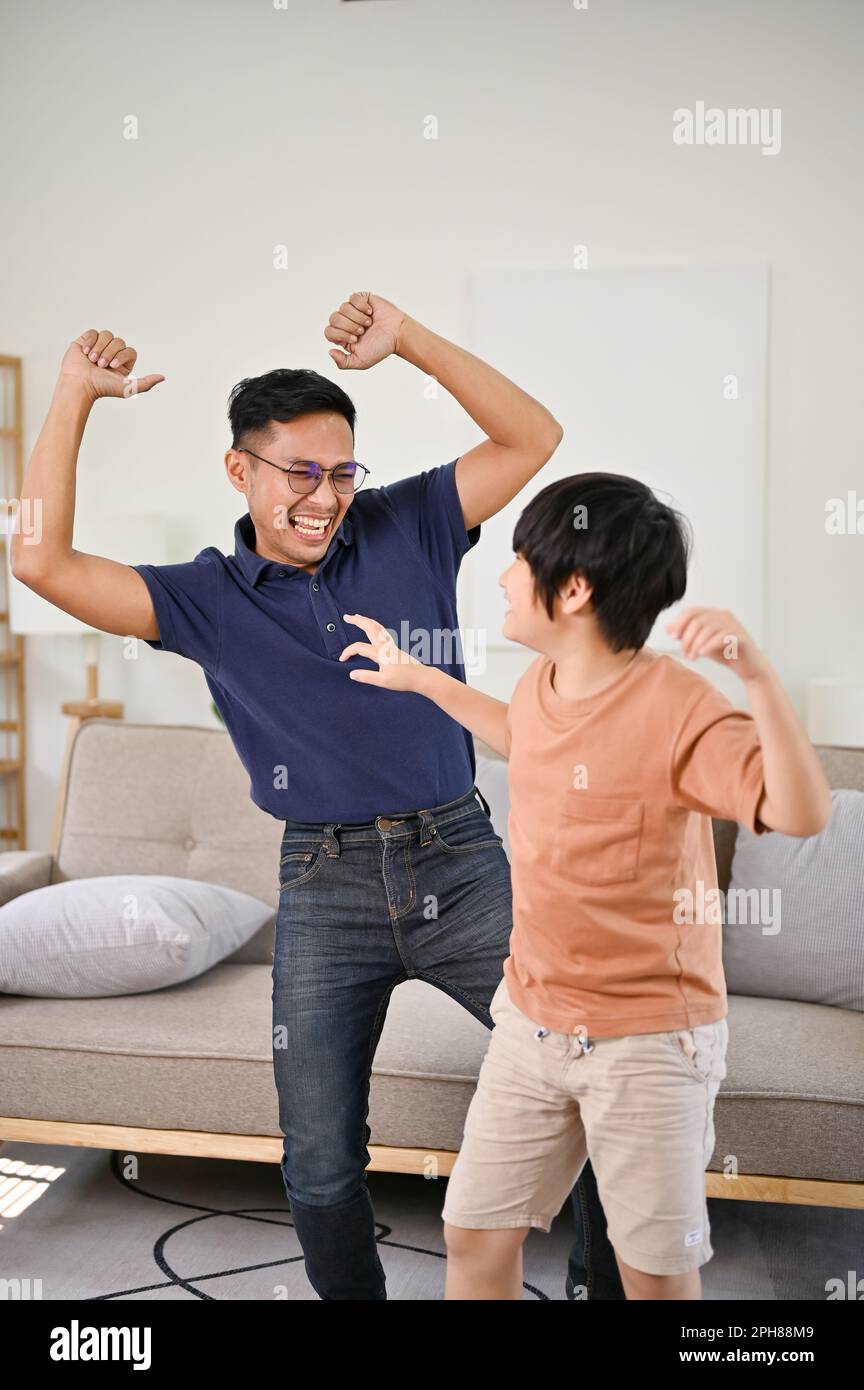 Cheerful and playful Asian dad and little son enjoying dancing in the living room together, having fun family time. happy family leisure concept Stock Photo