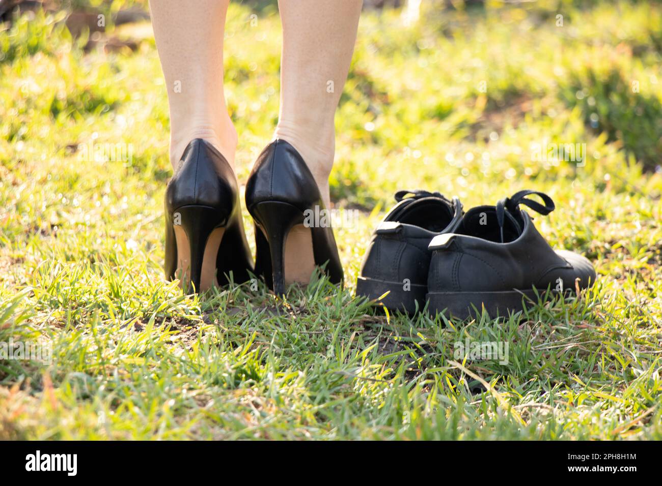 a girl in black leather high-heeled shoes and and next to it is another pair of black shoes on green grass in the sun Stock Photo