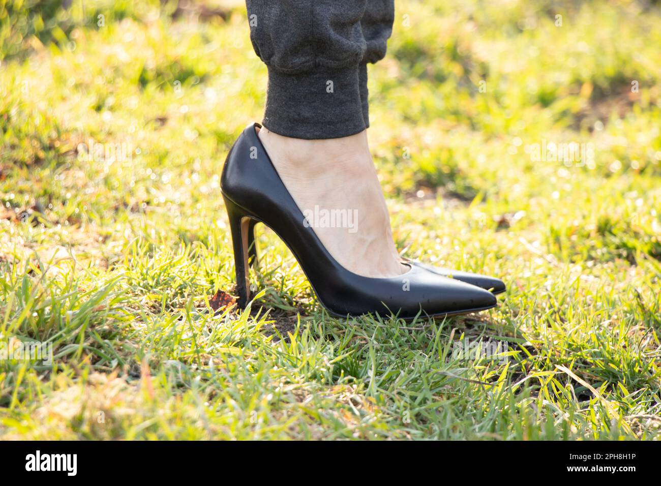 girl in black leather high-heeled shoes and gray sweatpants stands on green grass in the sun Stock Photo