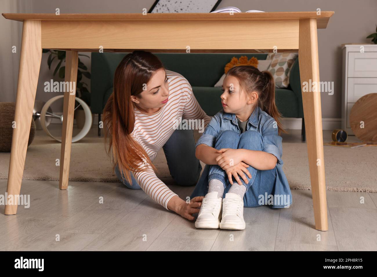 Scared mother with her little daughter hiding under table in living