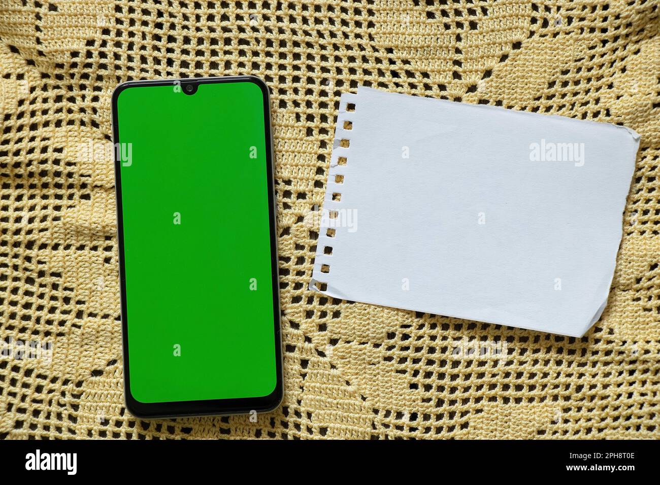 the phone with a green screen lies on a yellow lace napkin and next to a blank blank sheet of white paper on the table Stock Photo