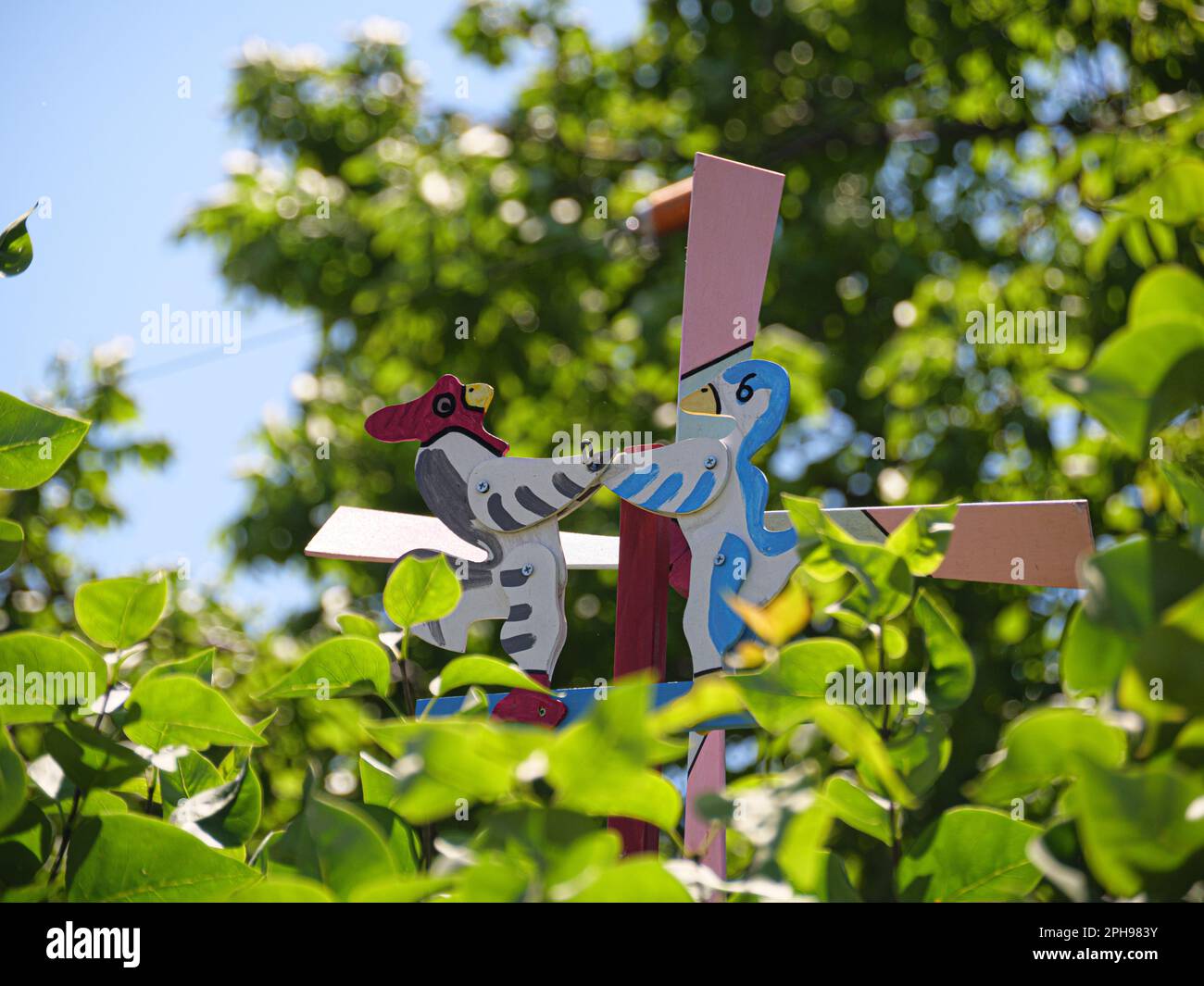 Colorful bird windmill decoration in the green outdoor garden forest at during the hot summer heatwave. War, red rooster, blue, yellow, white ducks Stock Photo