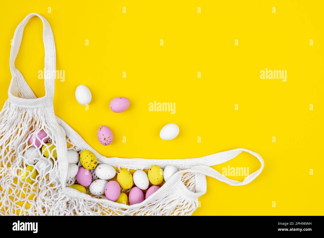 Colored Easter eggs in a mesh bag on a yellow background, flat lay. Stock Photo