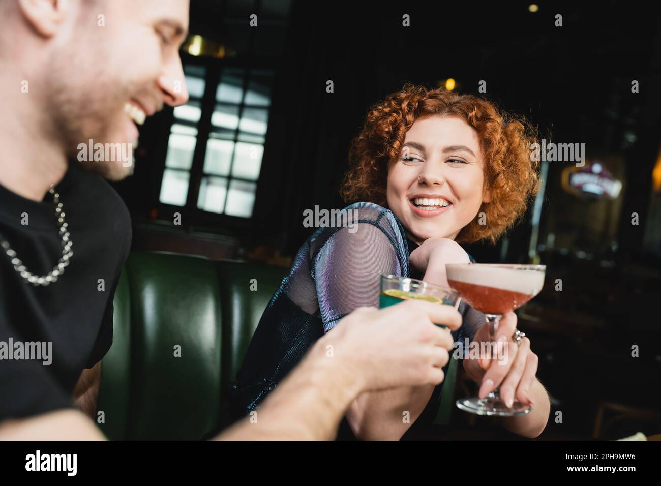Positive redhead woman clinking clover club cocktail with blurred friend in bar,stock image Stock Photo