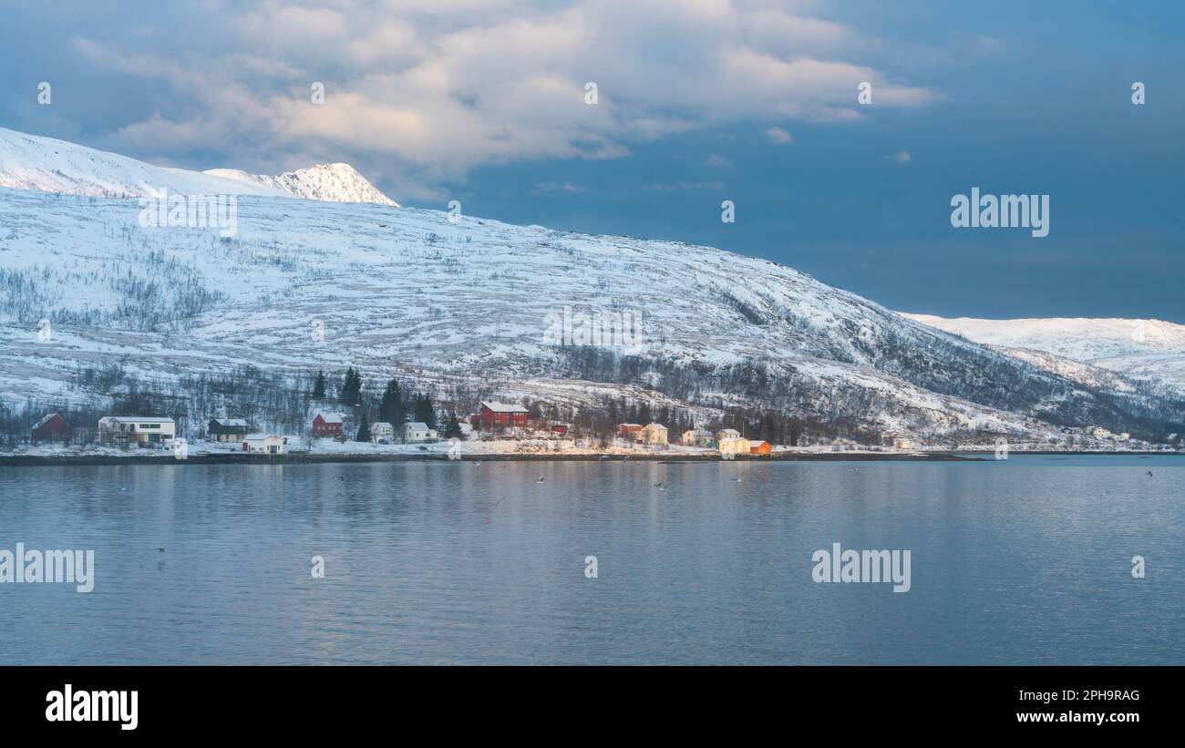 Morgenstimmung auf der Insel Senja und Kvaløya im Winter in Norwegen. Das Morgenrot färbt schneebedeckte Berge und Wolken rötlich, buntes Haus am Ufer Stock Photo