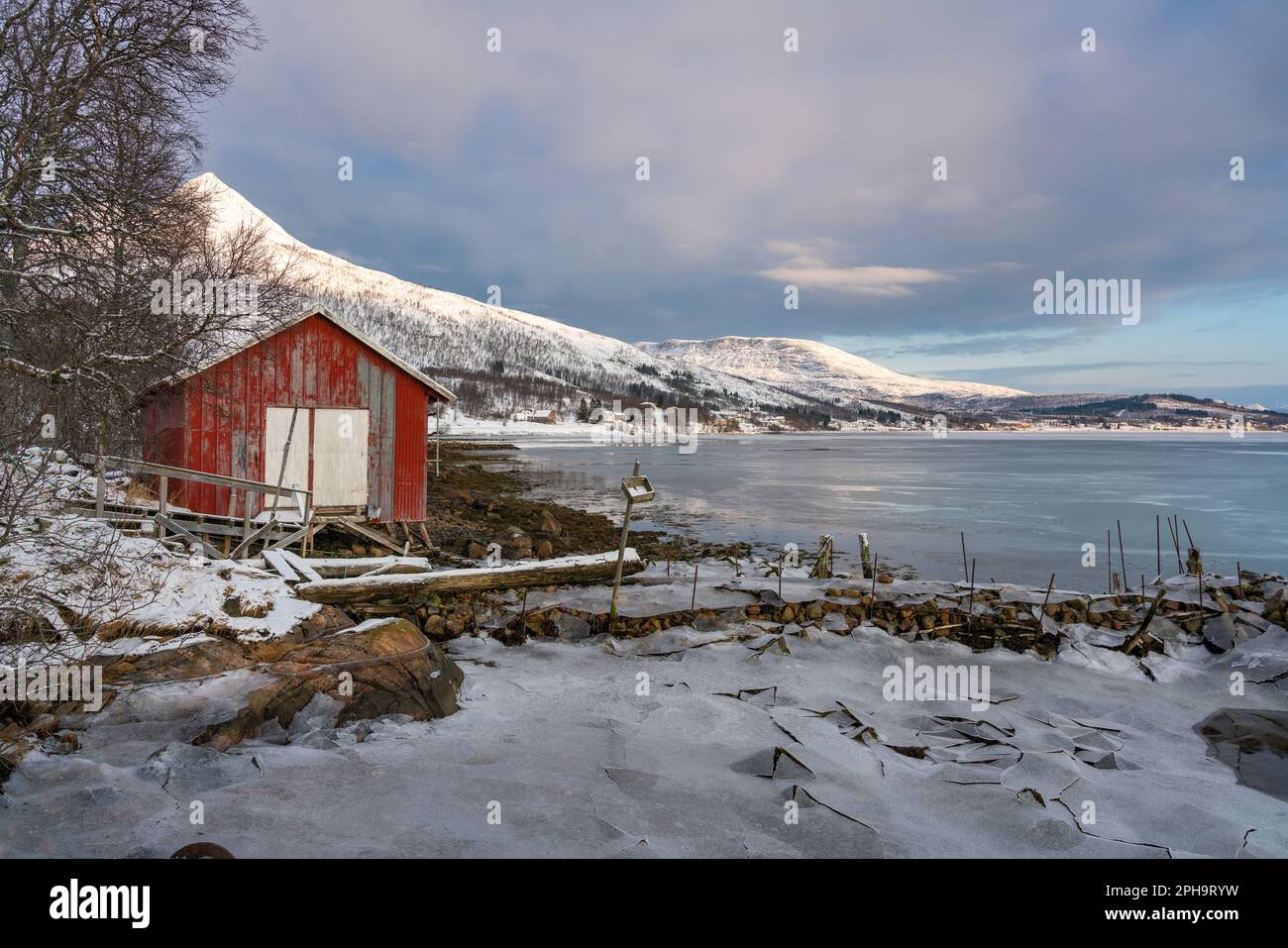 Morgenstimmung auf der Insel Senja und Kvaløya im Winter in Norwegen. Das Morgenrot färbt schneebedeckte Berge und Wolken rötlich, buntes Haus am Ufer Stock Photo