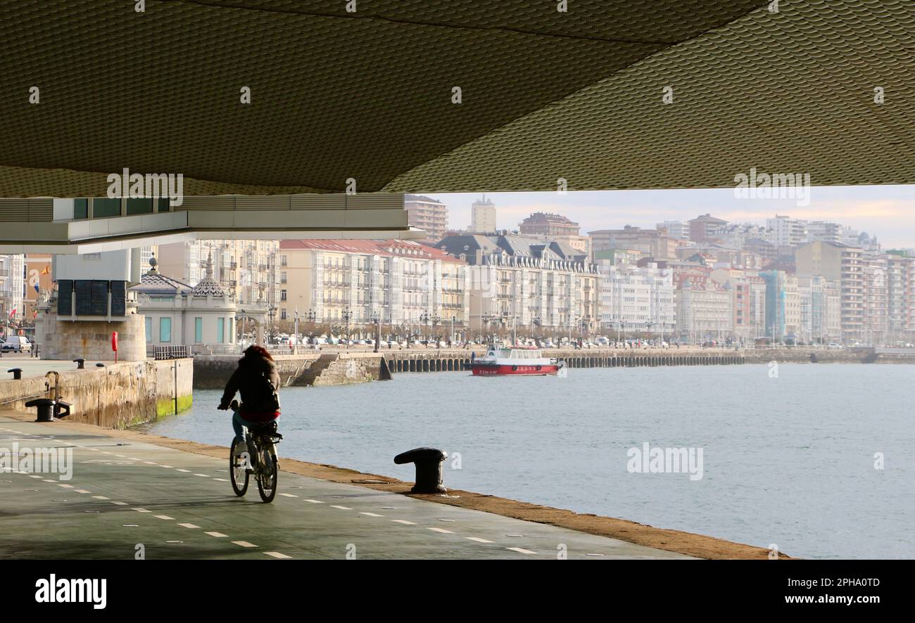 Cyclist passing under the Botin Centre next to the bay Santander Cantabria Spain a sunny spring morning with a local ferry arriving to the ferry stop Stock Photo