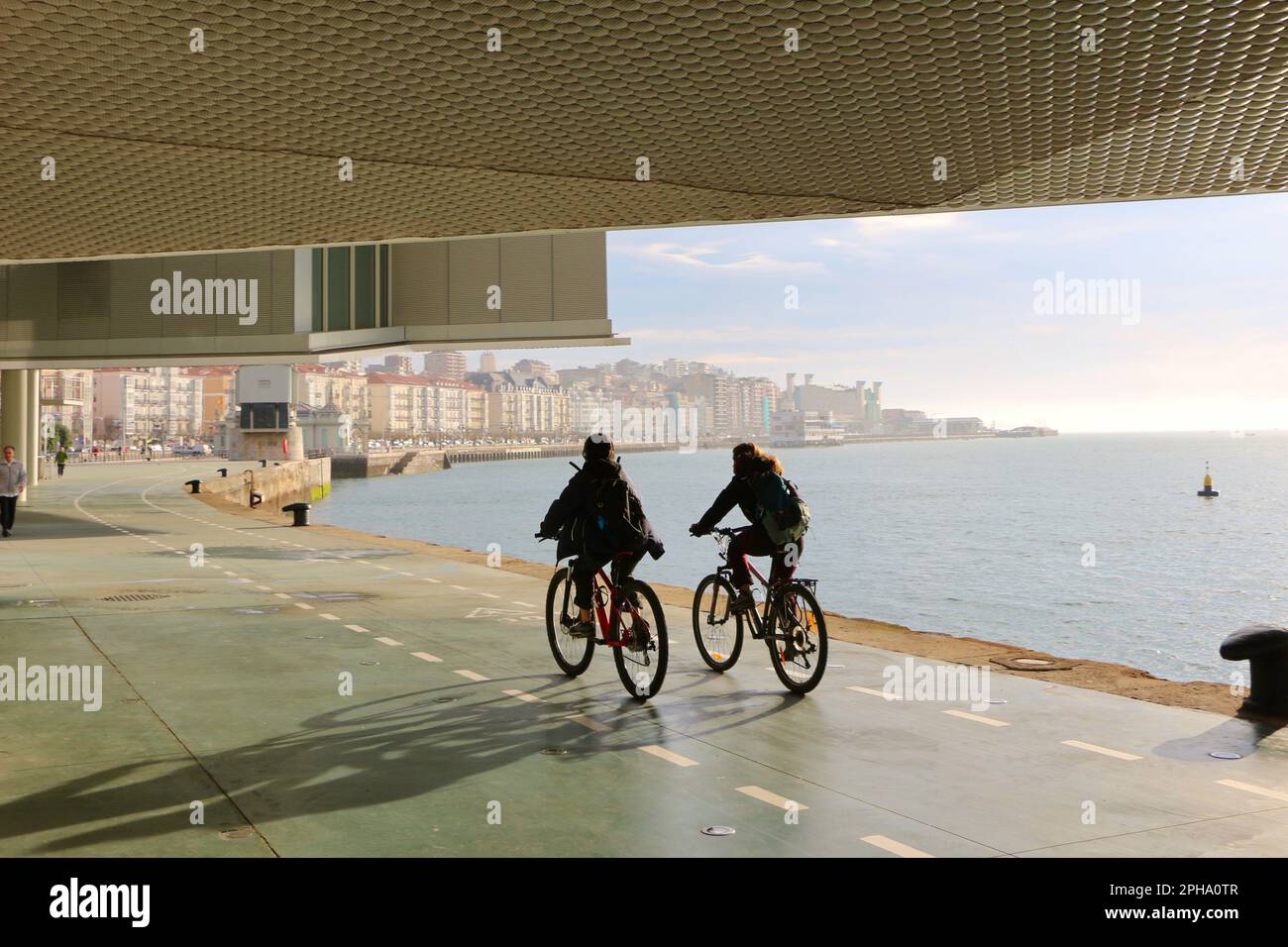 Two cyclists passing under the Botin Centre next to the bay Santander Cantabria Spain a sunny spring morning with the Festival Palace in the distance Stock Photo
