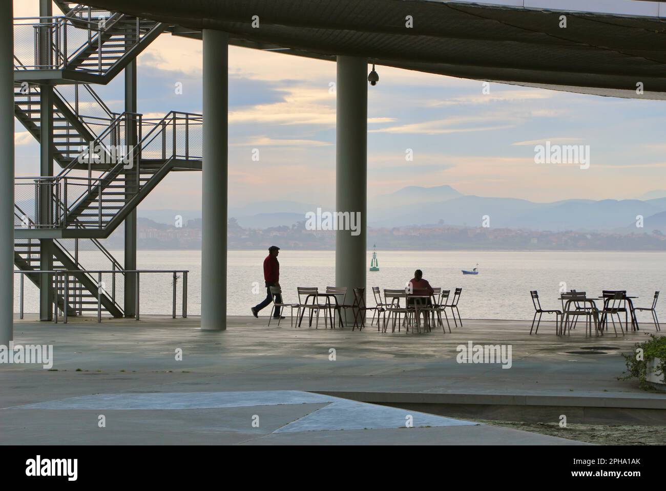 A man walking passing by a woman having breakfast at the open air terrace of the cafe Botin Arts Centre next to Santander Bay Cantabria Spain Stock Photo
