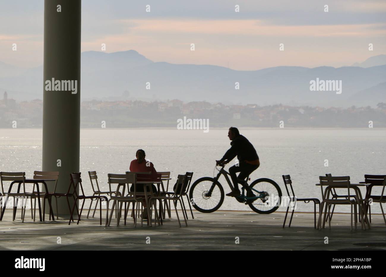 A cyclist passing by a woman having breakfast at the open air terrace of the cafe Botin Arts Centre next to Santander Bay Cantabria Spain Stock Photo
