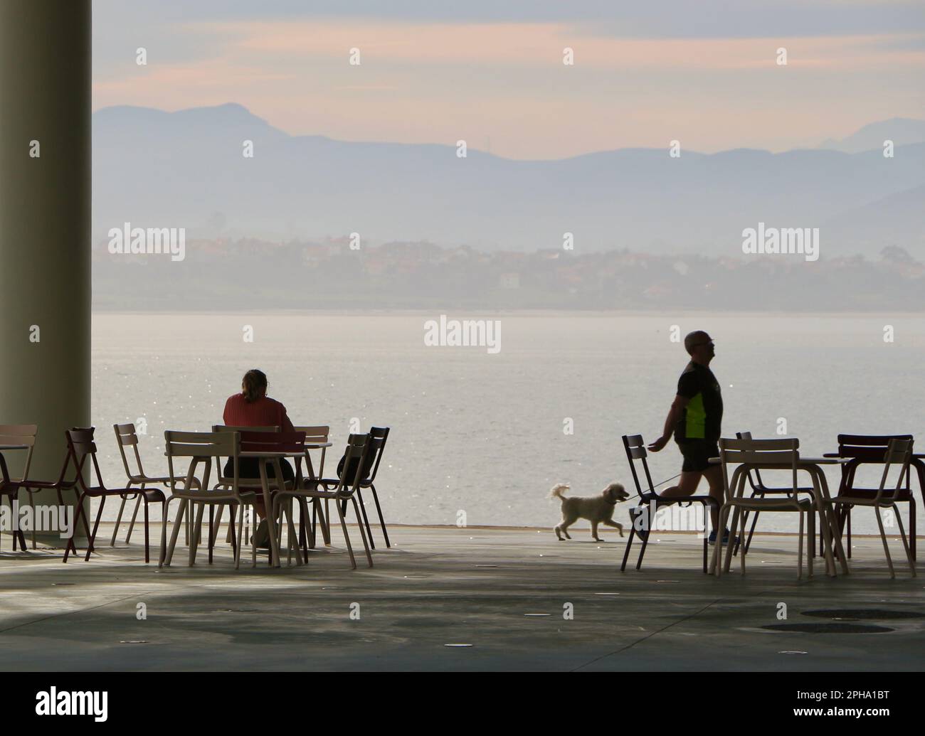 A man walking a dog passing by a woman having breakfast at the open air terrace of the cafe Botin Arts Centre next to Santander Bay Cantabria Spain Stock Photo