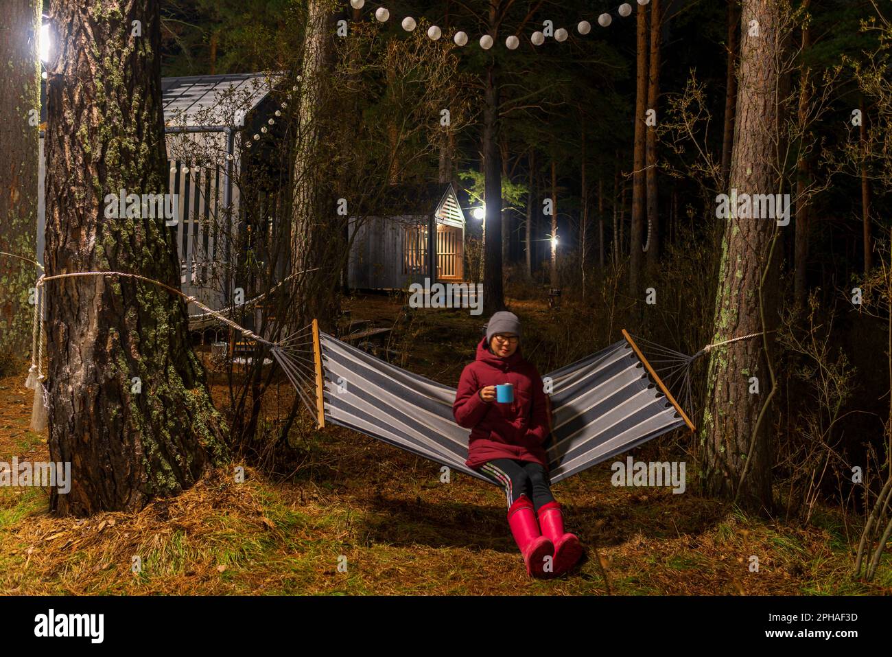 Altai, Russia - October 28, 2022 A girl in adidas pants sits on a hammock in autumn with a mug of tea in a jacket behind vacation houses. Stock Photo