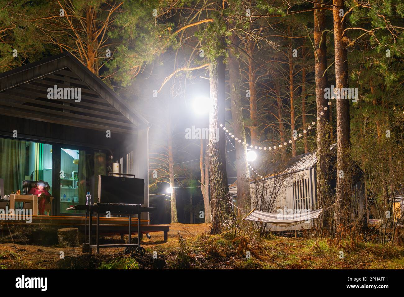 A brazier with fire and charcoal for a barbecue stands at night near a holiday cottage with a woman on a chair with lantern lights and garlands in Alt Stock Photo