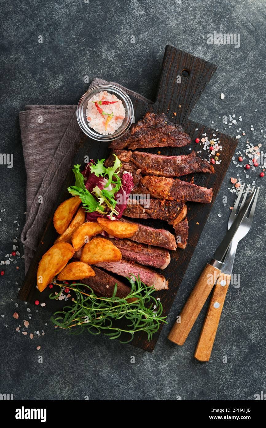 Steaks. Sliced grilled meat steak New York, Ribeye or Chuck roll with with garnished with salad and french fries on black marble board on old wooden b Stock Photo