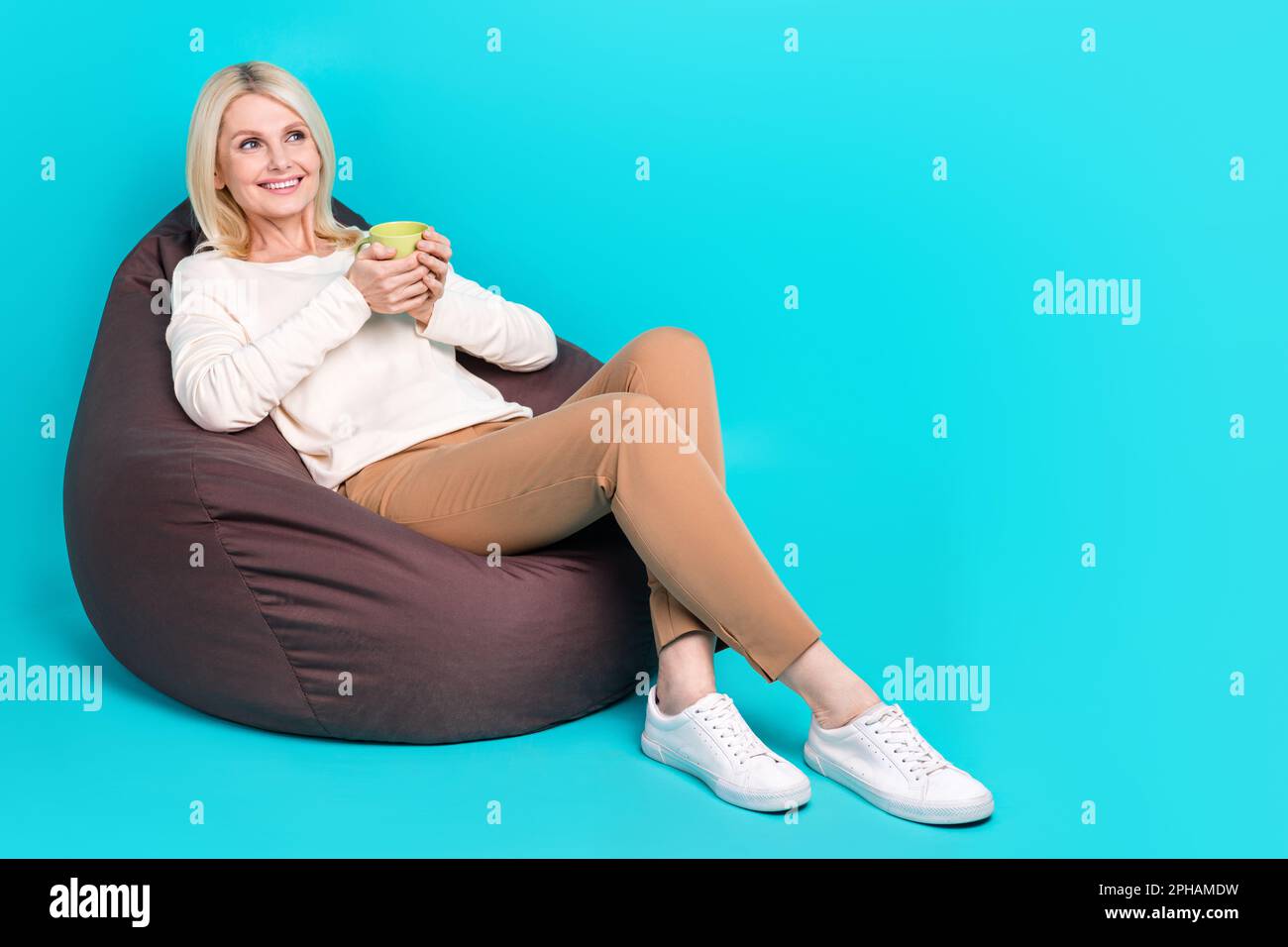 Full length photo of dreaming lady pensioner pouf hold aroma tasty drink tea look empty space sit beanbag dorm room isolated on cyan color background Stock Photo