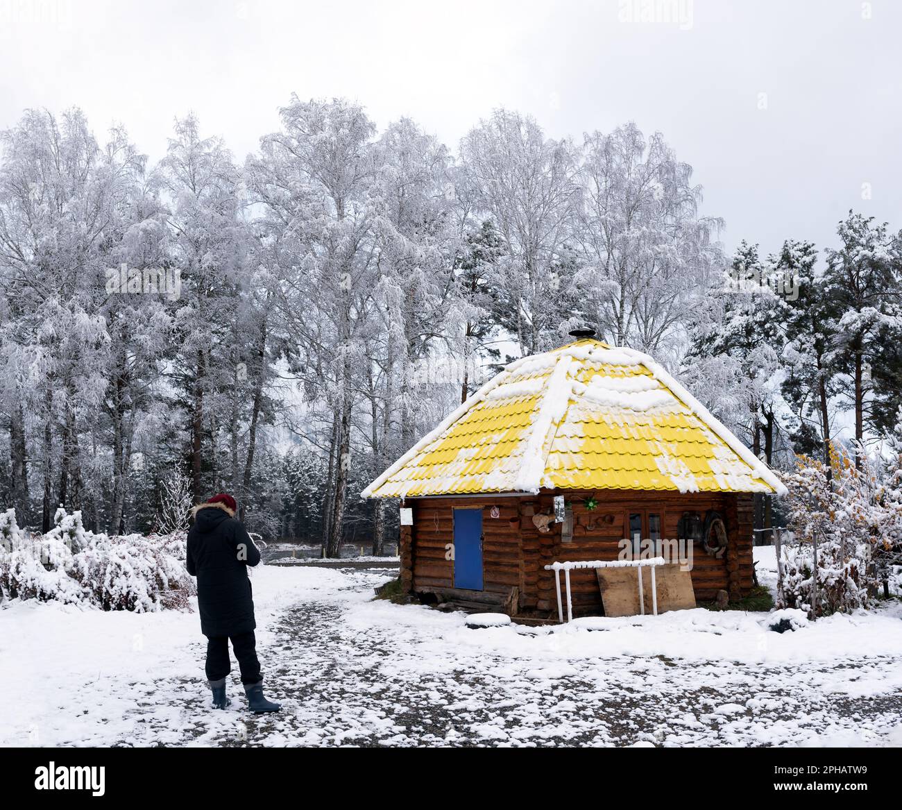 An elderly woman stands at the round house of the ail under the trees in the snow in winter in the Altai in Siberia. Stock Photo