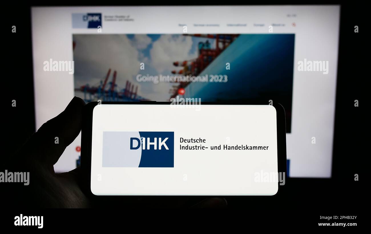 Person holding smartphone with logo of Deutsche Industrie- und Handelskammer (DIHK) on screen in front of website. Focus on phone display. Stock Photo