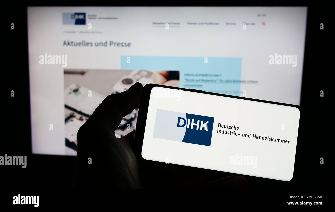 Person holding cellphone with logo of Deutsche Industrie- und Handelskammer (DIHK) on screen in front of webpage. Focus on phone display. Stock Photo