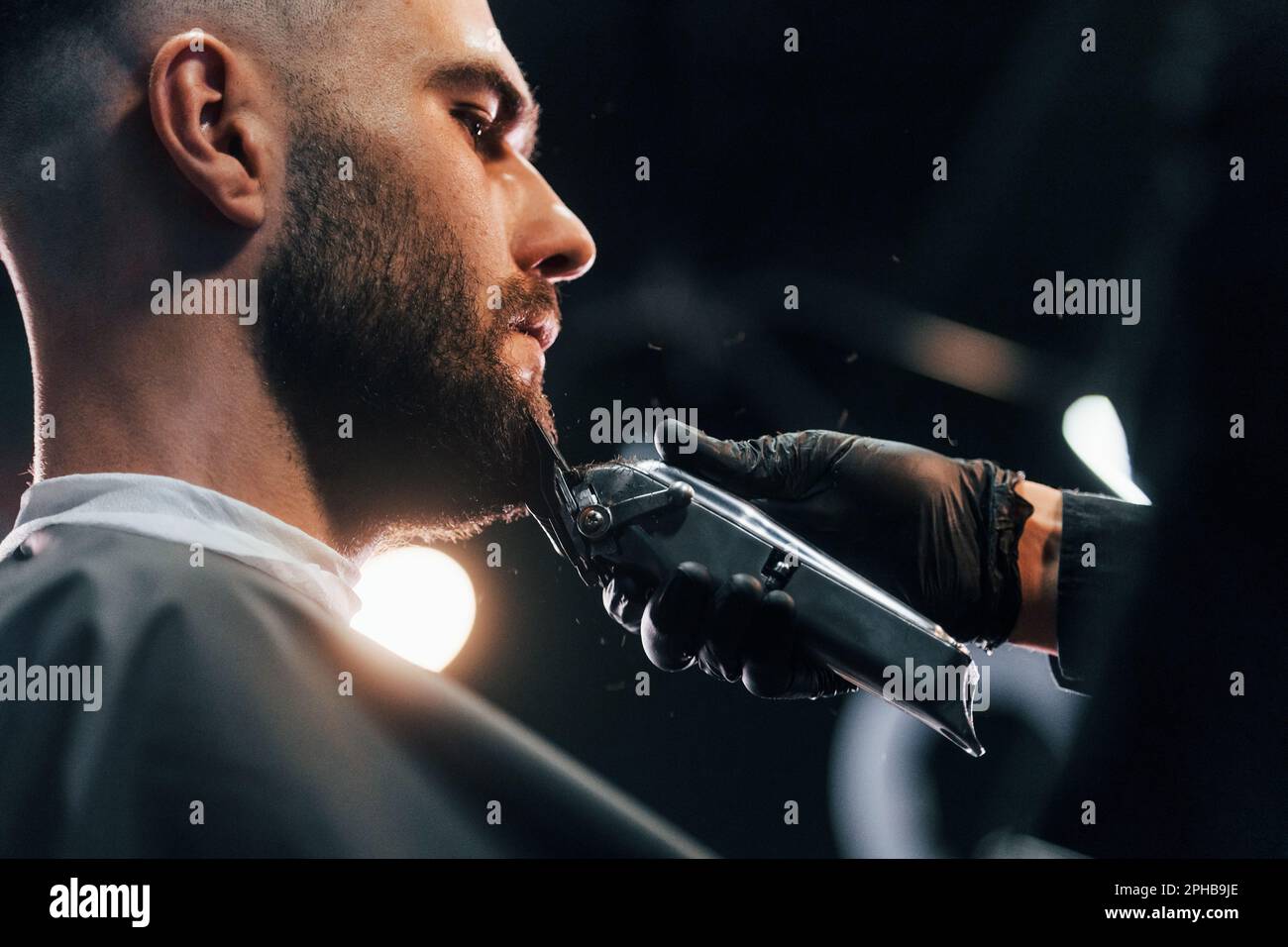 Young man with stylish hairstyle sitting and getting his beard shaved