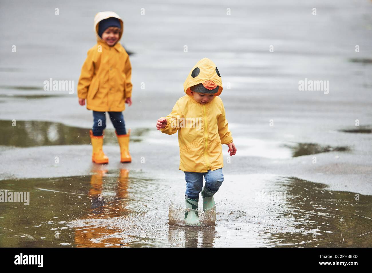 Two kids in yellow waterproof cloaks and boots playing outdoors after the rain together Stock