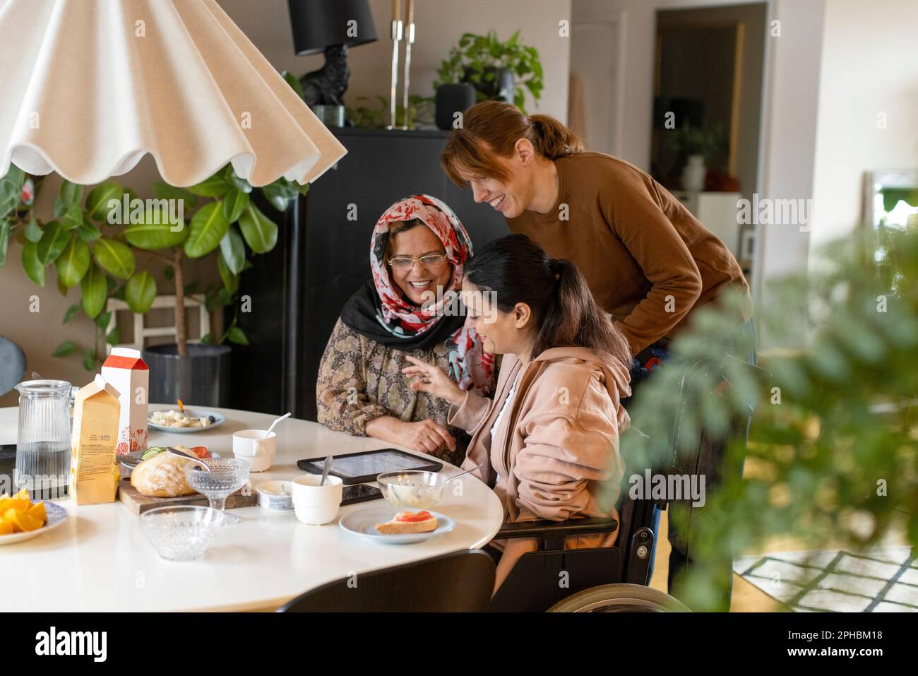 Female caregiver talking to woman with paraplegia and her mother sitting at dining table in home Stock Photo