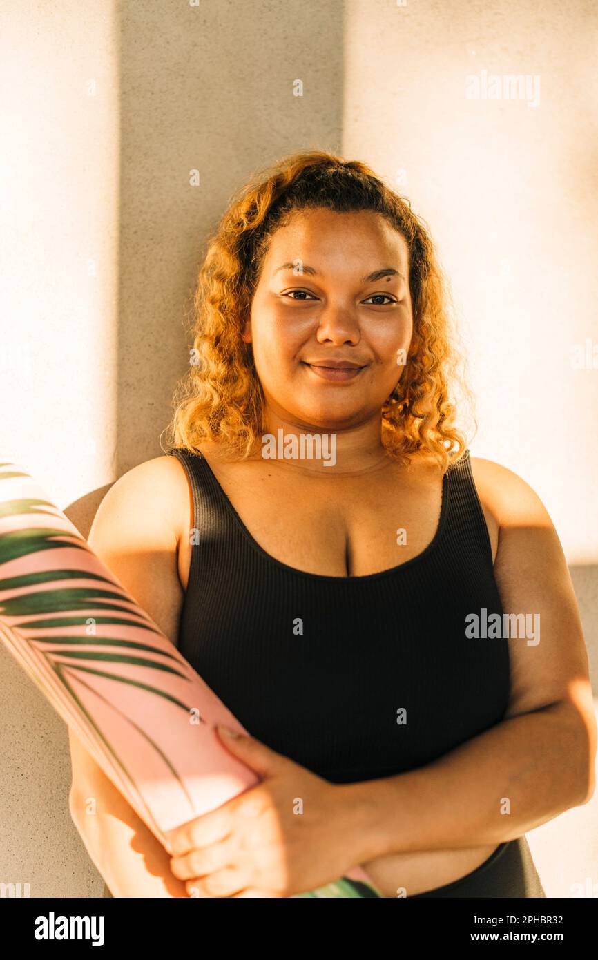 Portrait of voluptuous woman holding exercise mat against wall Stock