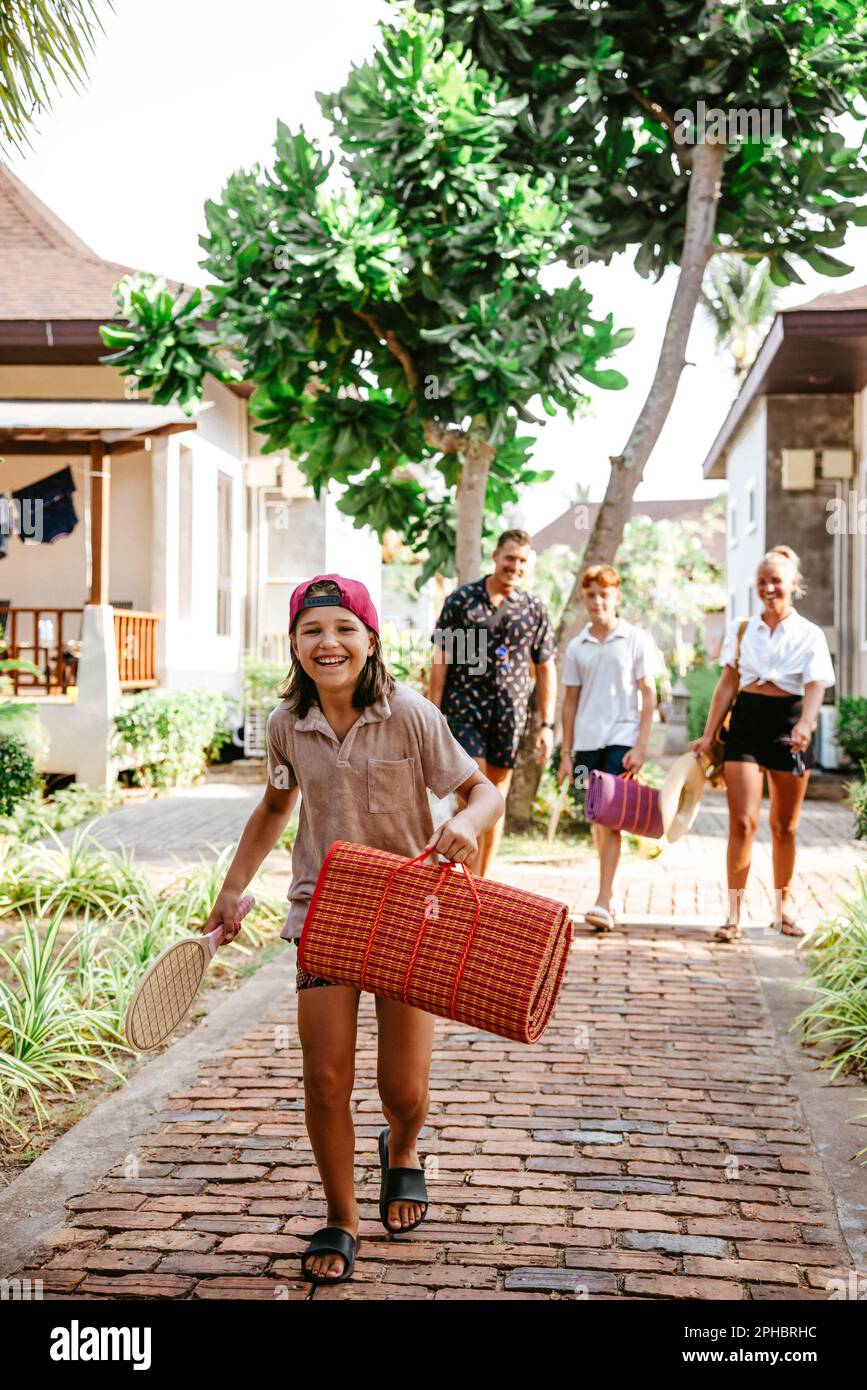 Happy girl holding mat while walking on footpath with family at tourist resort Stock Photo