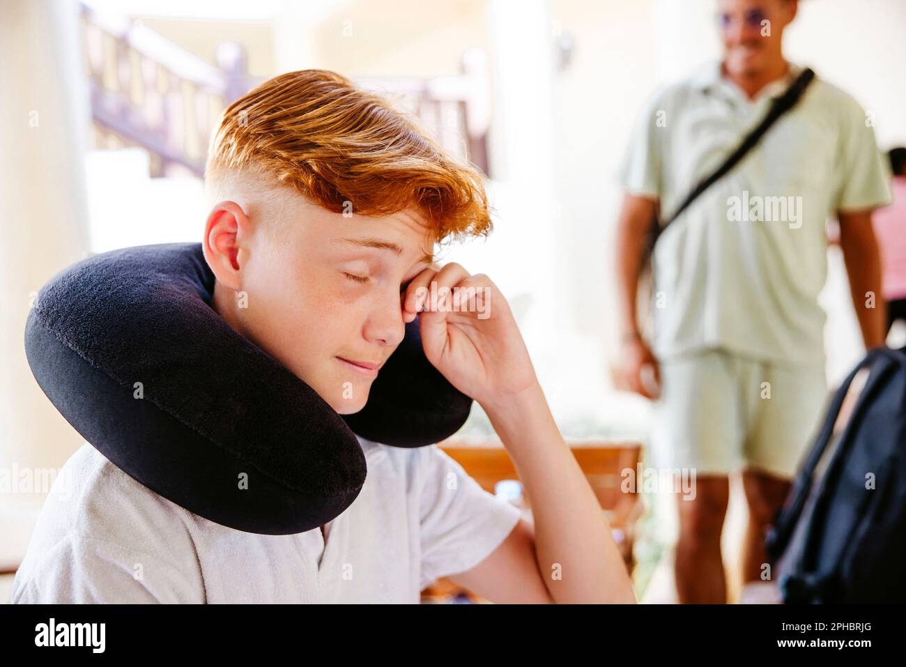 Tired boy with neck pillow rubbing eye in villa during vacation Stock Photo