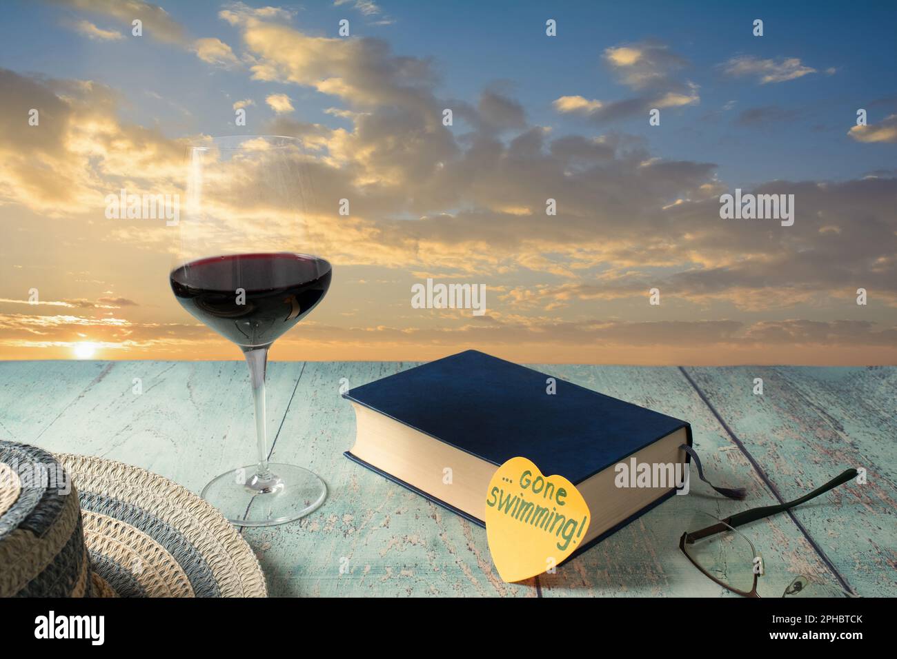 Gone swimming! A glass of red wine, a good book, a straw hat, a beautiful sunset, and a post-it note. The call of adventure and holiday pleasures. Stock Photo