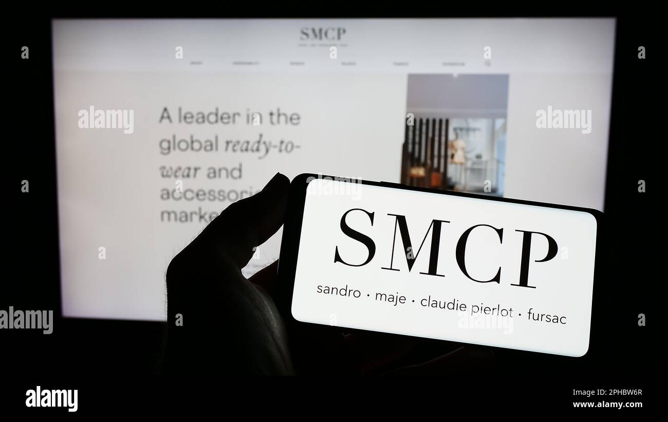 Person holding mobile phone with logo of French luxury fashion company SMCP SA on screen in front of busness web page. Focus on phone display. Stock Photo