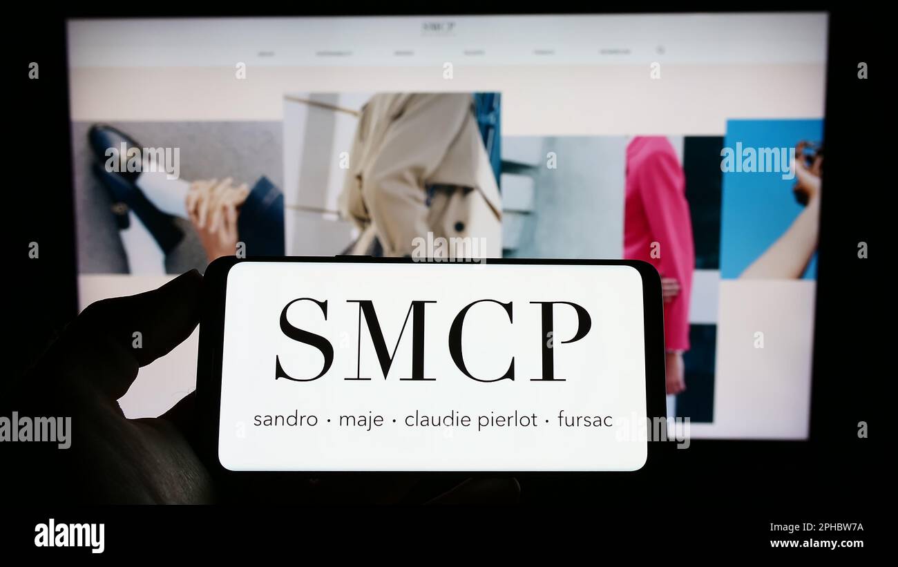 Person holding cellphone with logo of French luxury fashion company SMCP S.A. on screen in front of business webpage. Focus on phone display. Stock Photo