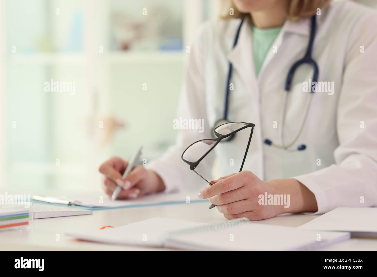 Doctor writes notes holding glasses at table in hospital Stock Photo