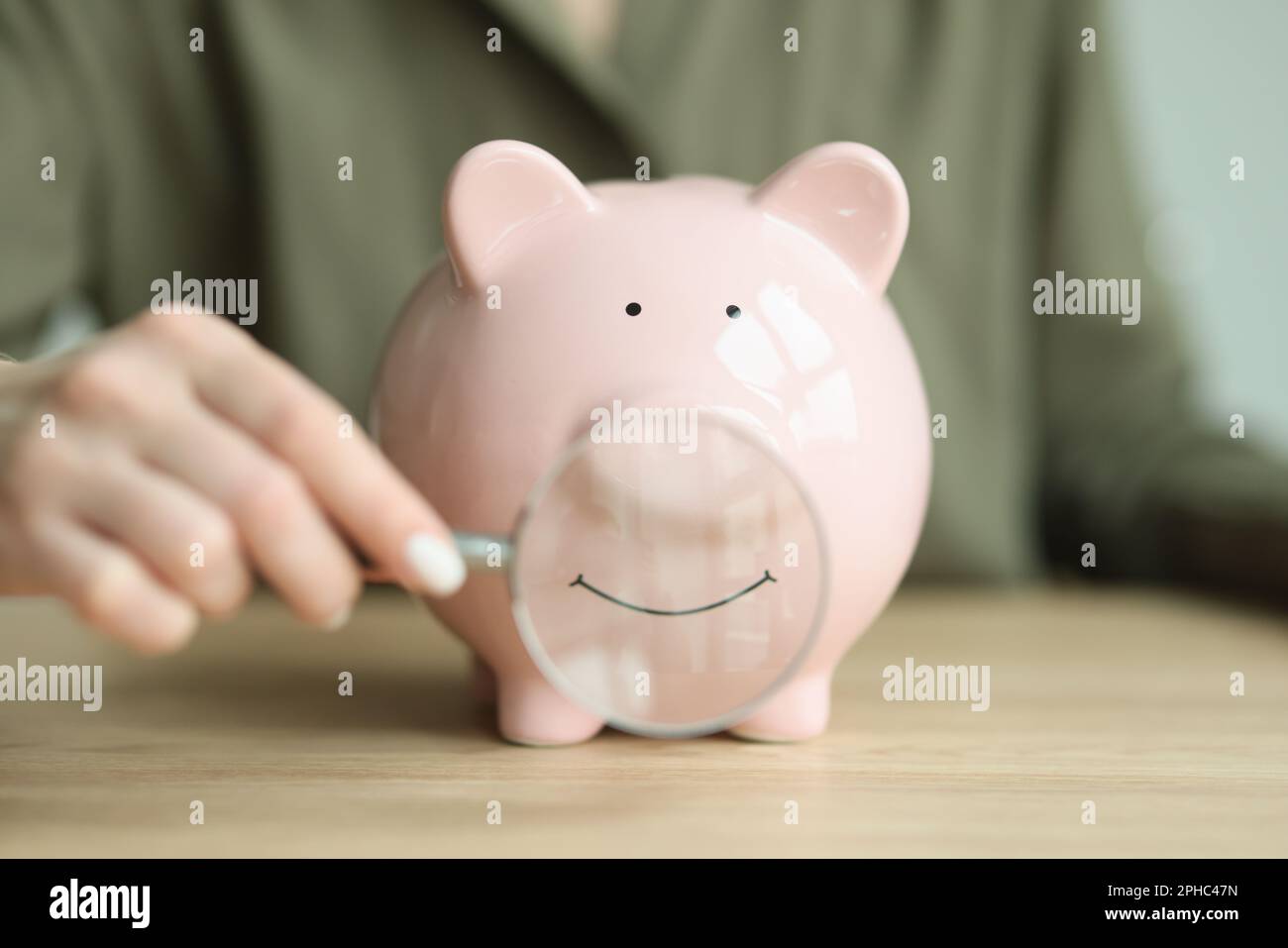 Hand of woman putting magnifier glass to mouth of piggy bank Stock Photo