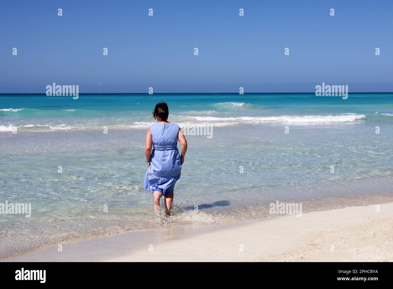 Woman in blue dress running in azure sea water. Travel and beach vacation on sandy coast Stock Photo