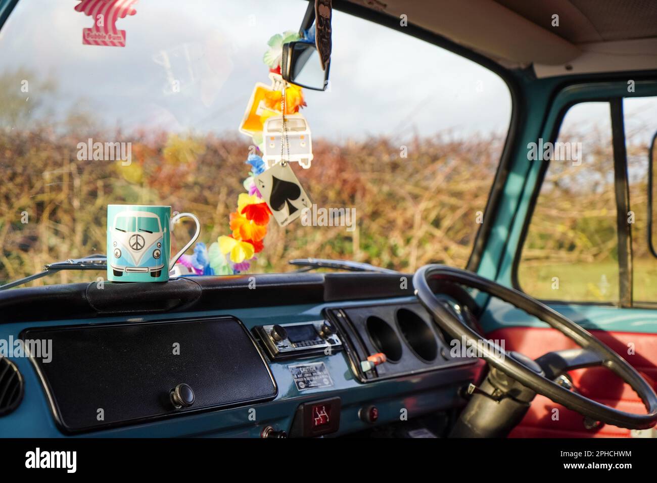 Looking out over the dashboard and steering wheel of a 'Bay Window' VW camper van, with a coffee mug and garland of brightly coloured flowers hanging Stock Photo