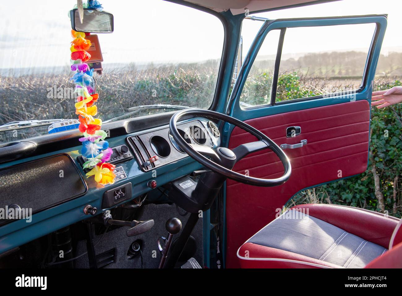 Looking out over the dashboard and steering wheel of a 'Bay Window' VW camper van over fields, with a garland of brightly coloured flowers hanging Stock Photo