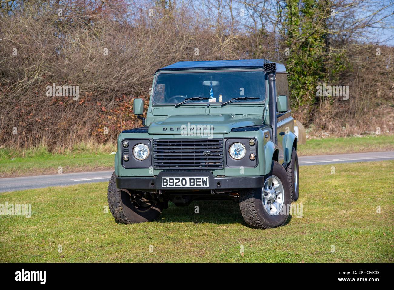 Green Land Rover Defender equipped with five-spoke alloy wheels and snorkel, parked on grass with a country lane and trees behind Stock Photo