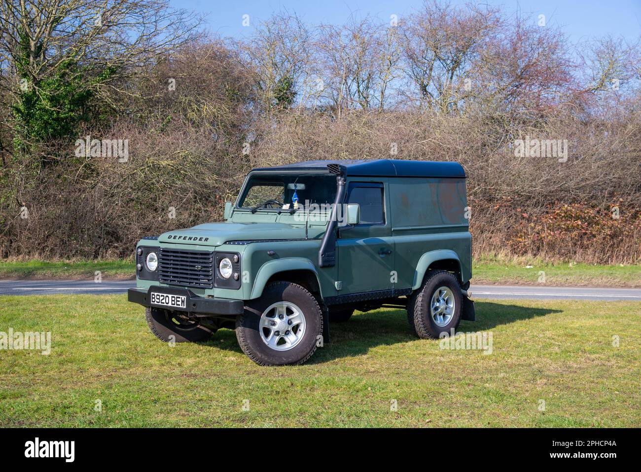 Green Land Rover Defender equipped with five-spoke alloy wheels and snorkel, parked on grass with a country lane and trees behind Stock Photo