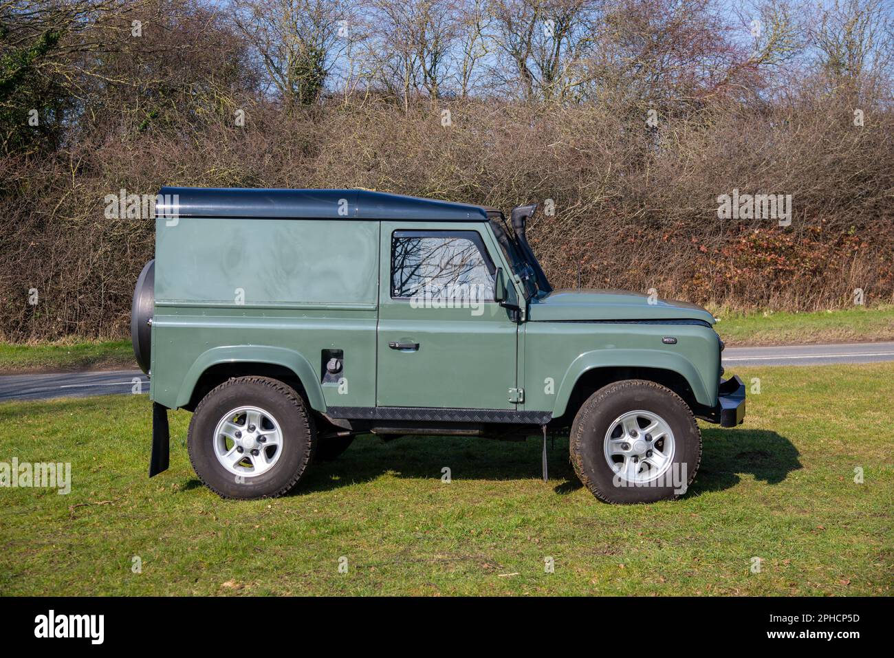 Green Land Rover Defender equipped with five-spoke alloy wheels and snorkel, parked on grass with a country lane and trees behind Stock Photo