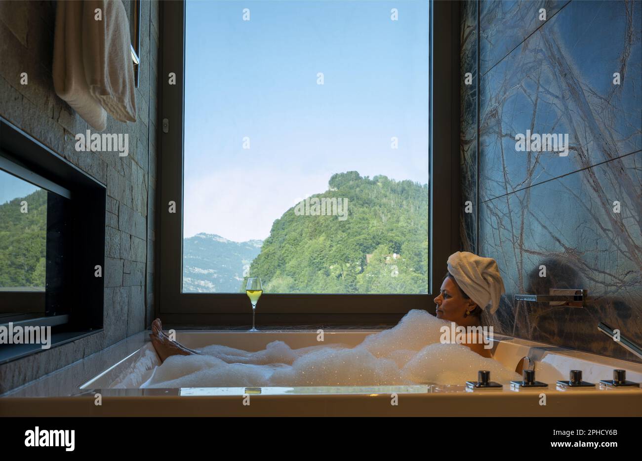 Woman Lying in a Bathtub with a Glass of Champagne and Enjoy the Window View over Mountain in a Sunny Summer Day in Burgenstock, Nidwalden, Switzerlan Stock Photo